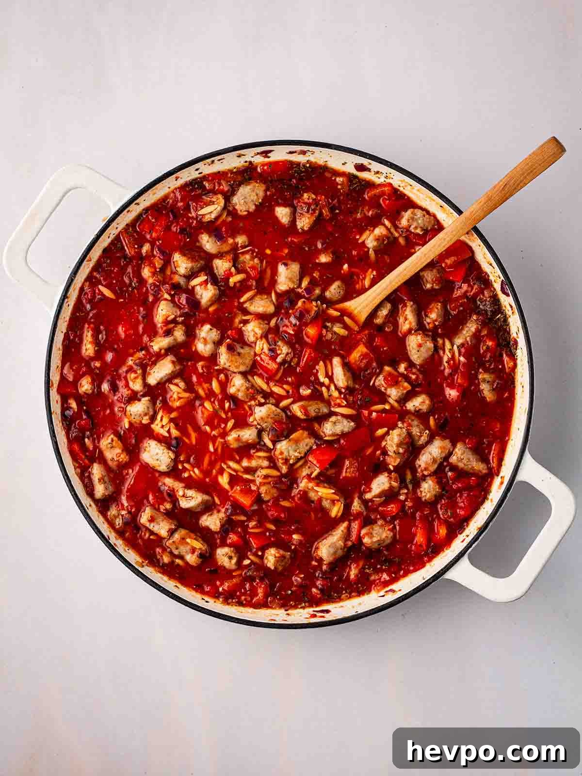 One-Pot Sausage and Orzo Dinner 7 Chunks of sausage and red pepper in a pan with orzo and tomato sauce, with a wooden spoon.