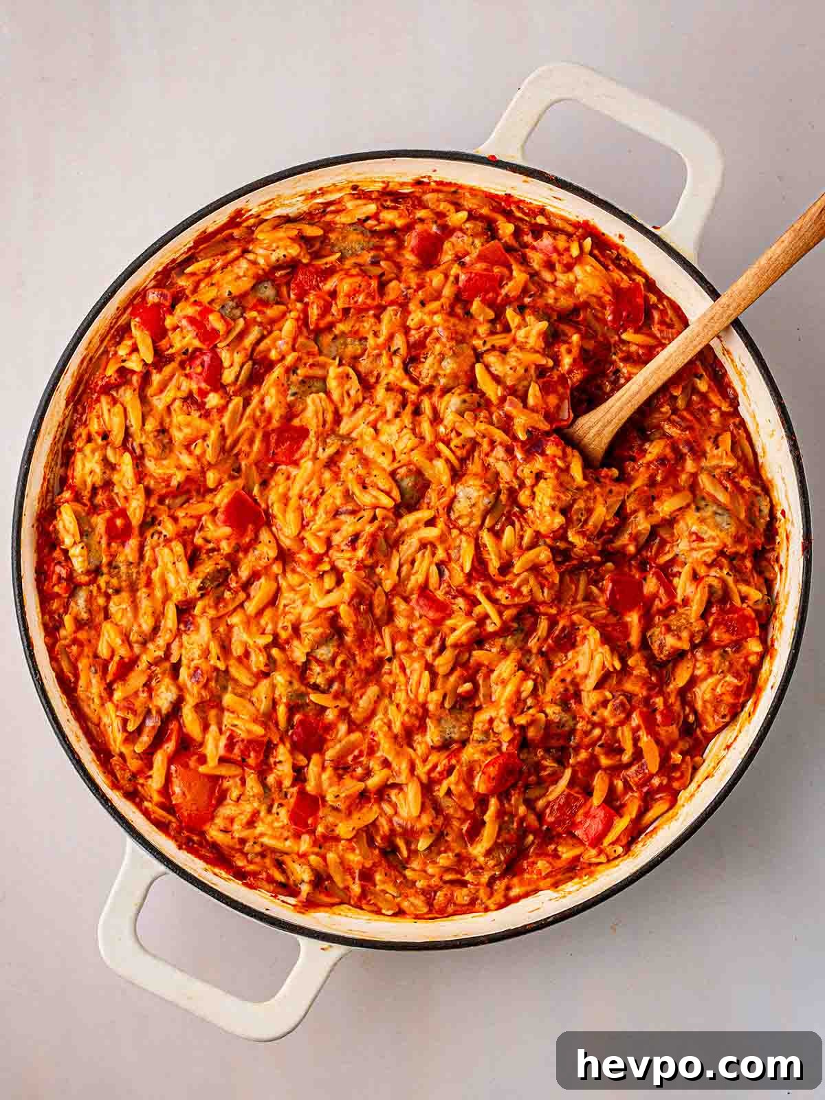 One-Pot Sausage and Orzo Dinner 8 A spoon stirring a white pan filled with Sausage Orzo for for step 4 in the recipe for Sausage Orzo.