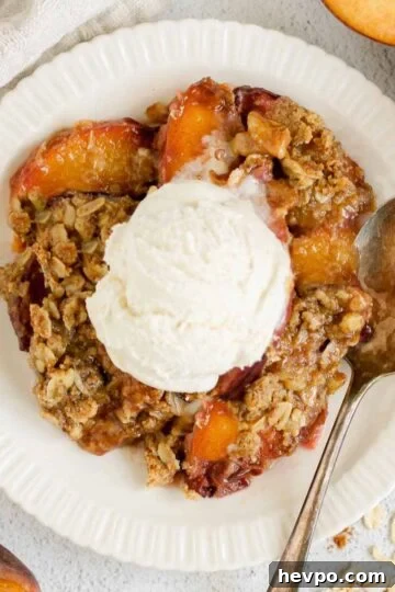 Serving of peach crisp on a round plate topped with ice cream and a vintage spoon on the plate with sliced peaches on the sides of the plate.