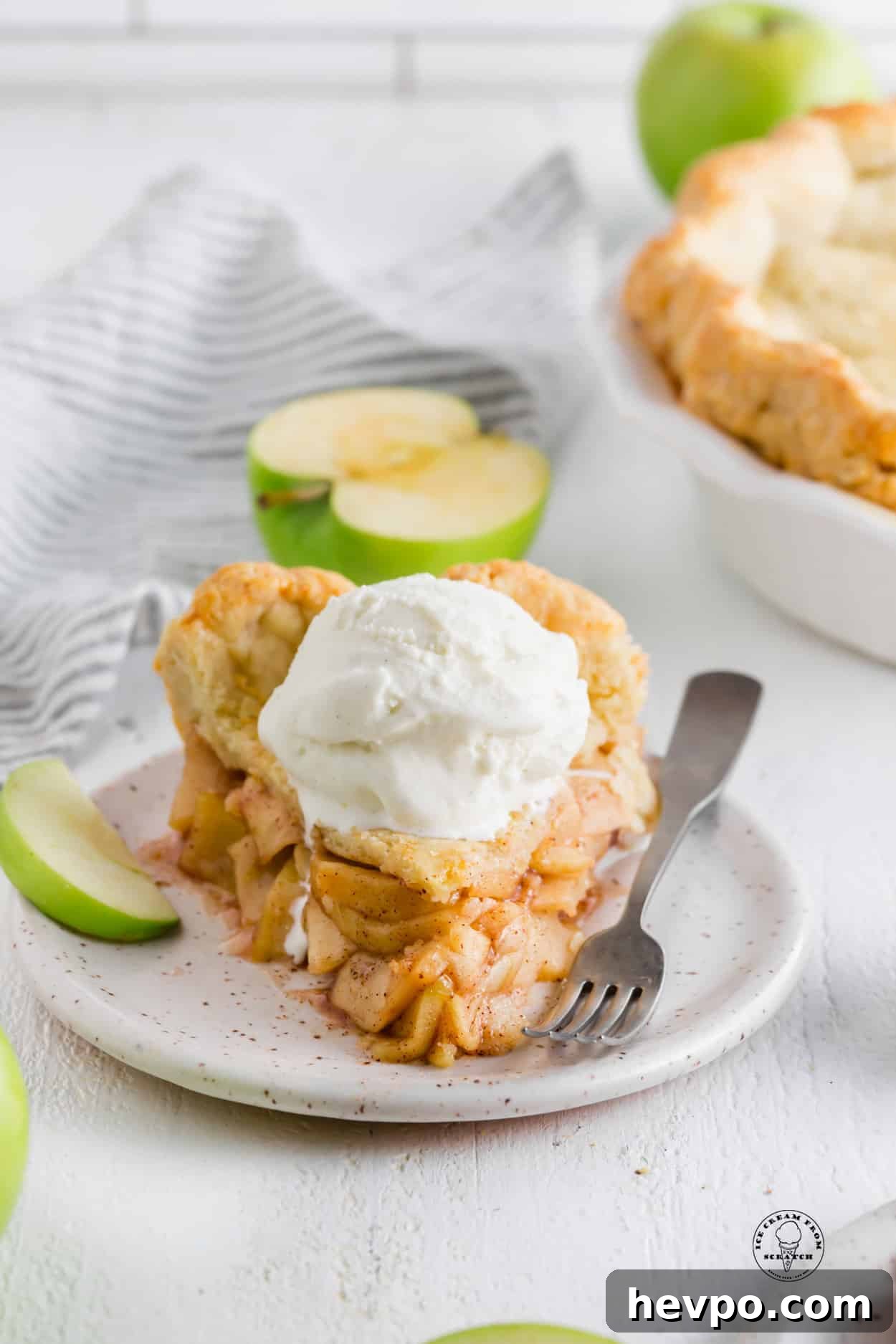 Apple Pie à la Mode: Classic Warm Apple Pie with Ice Cream 2 a thick slice of homemade apple pie with a scoop of vanilla ice cream on top. A fork and a slice of green apple are on the plate.