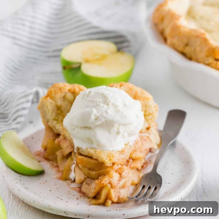 a thick slice of homemade apple pie with a scoop of vanilla ice cream on top. A fork and a slice of green apple are on the plate.
