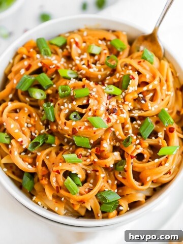 A white bowl filled with creamy peanut butter noodles. They are garnished with green onions and white sesame seeds.