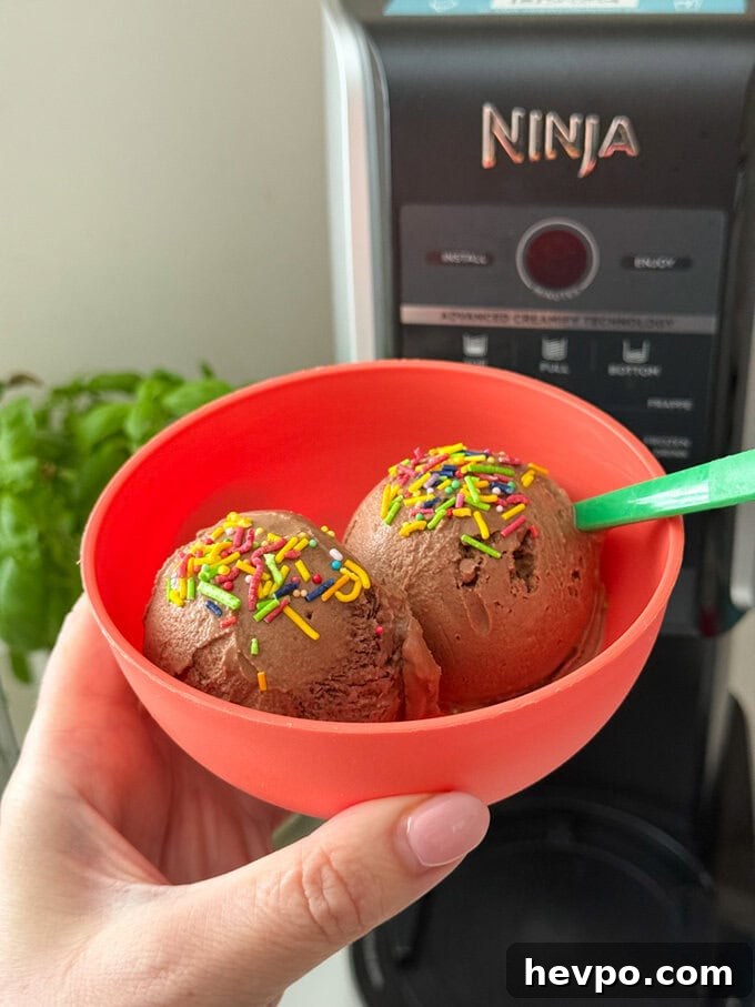 Delicious chocolate flavoured frozen yogurt made with just three simple ingredients, served in a bright red bowl with a green plastic spoon, and bright sprinkles over the scooped yogurt.