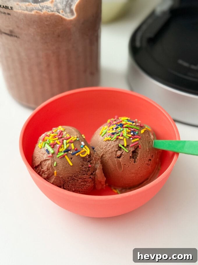 Delicious chocolate flavoured frozen yogurt made with just three simple ingredients, served in a bright red bowl with a green plastic spoon, and bright sprinkles over the scooped yogurt.