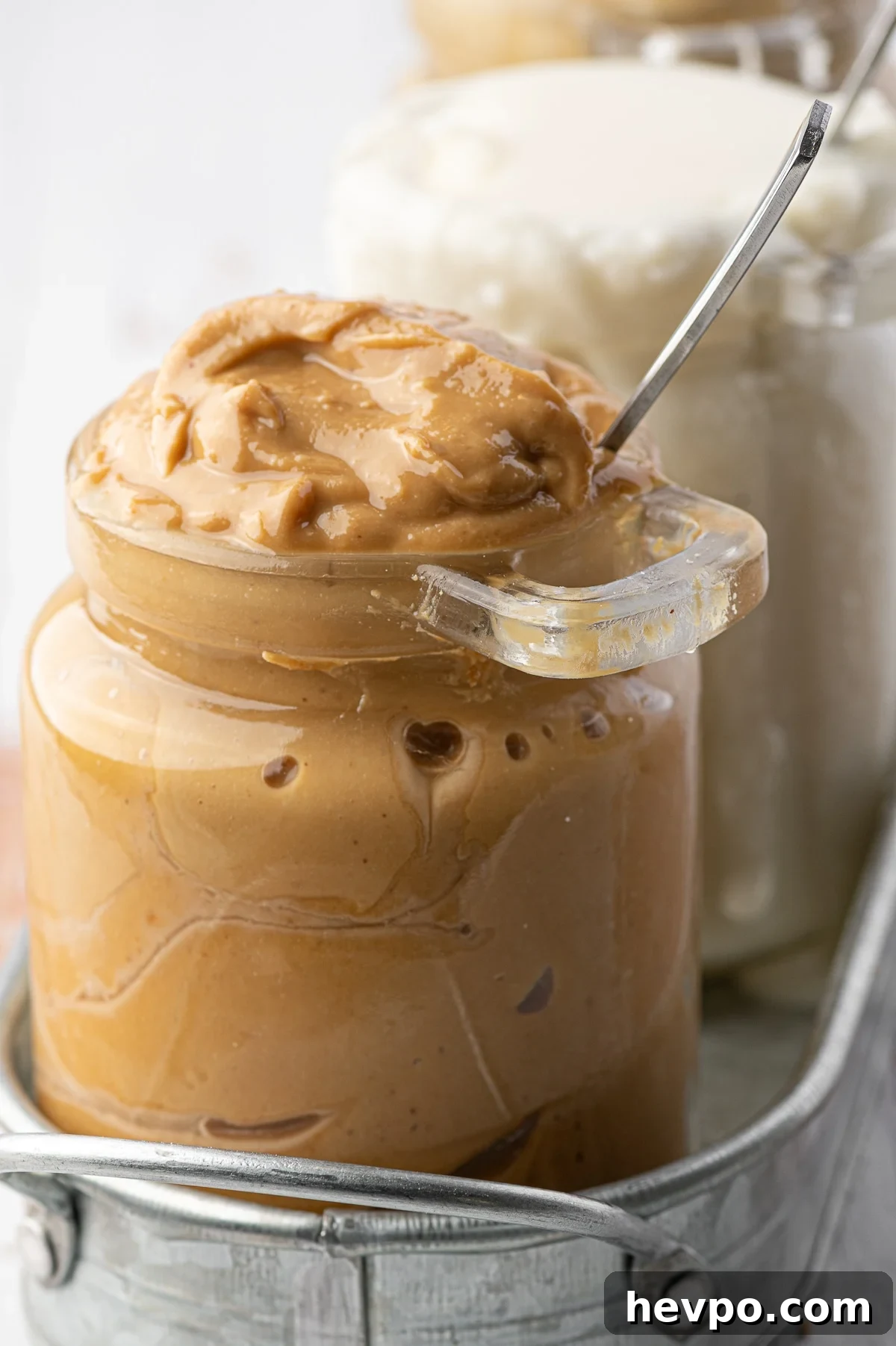An overfull glass jar of homemade natural peanut butter with a metal spoon stuck in the jar. 