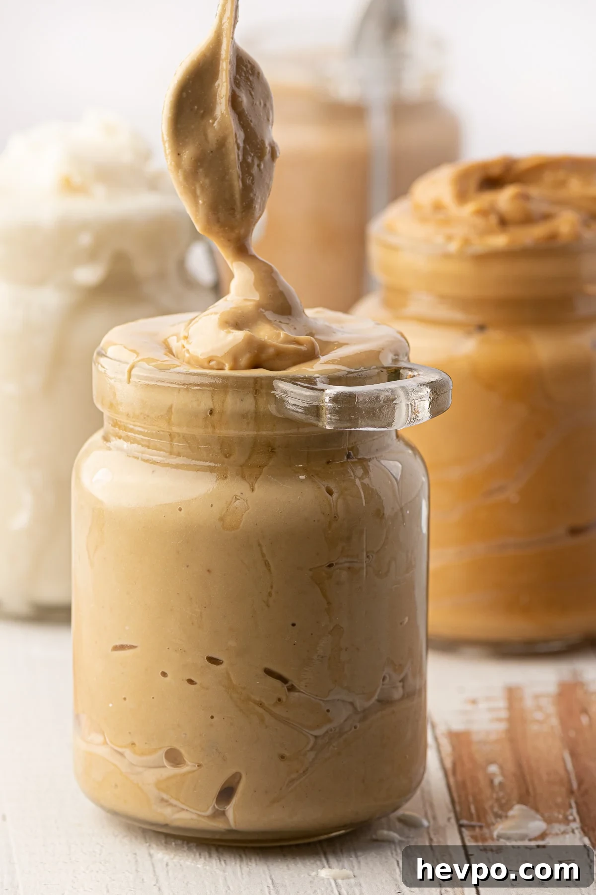 A jar of cashew butter being spooned into a glass jar with jars of overfull nut butters in the background 