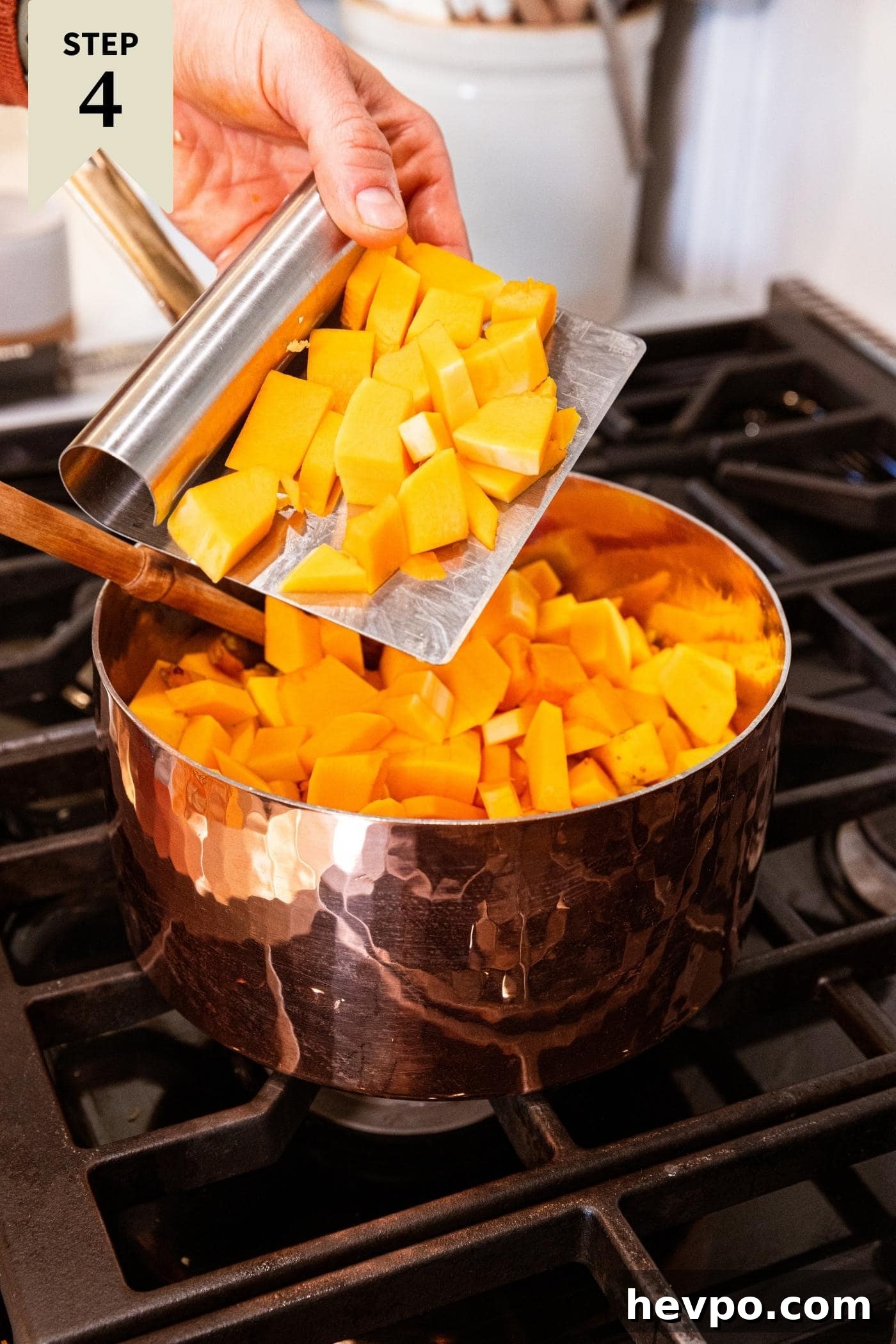 Hand using metal bench scraper to put chunks of chopped butternut squash into copper pot on stovetop already filled with carrots and onions.
