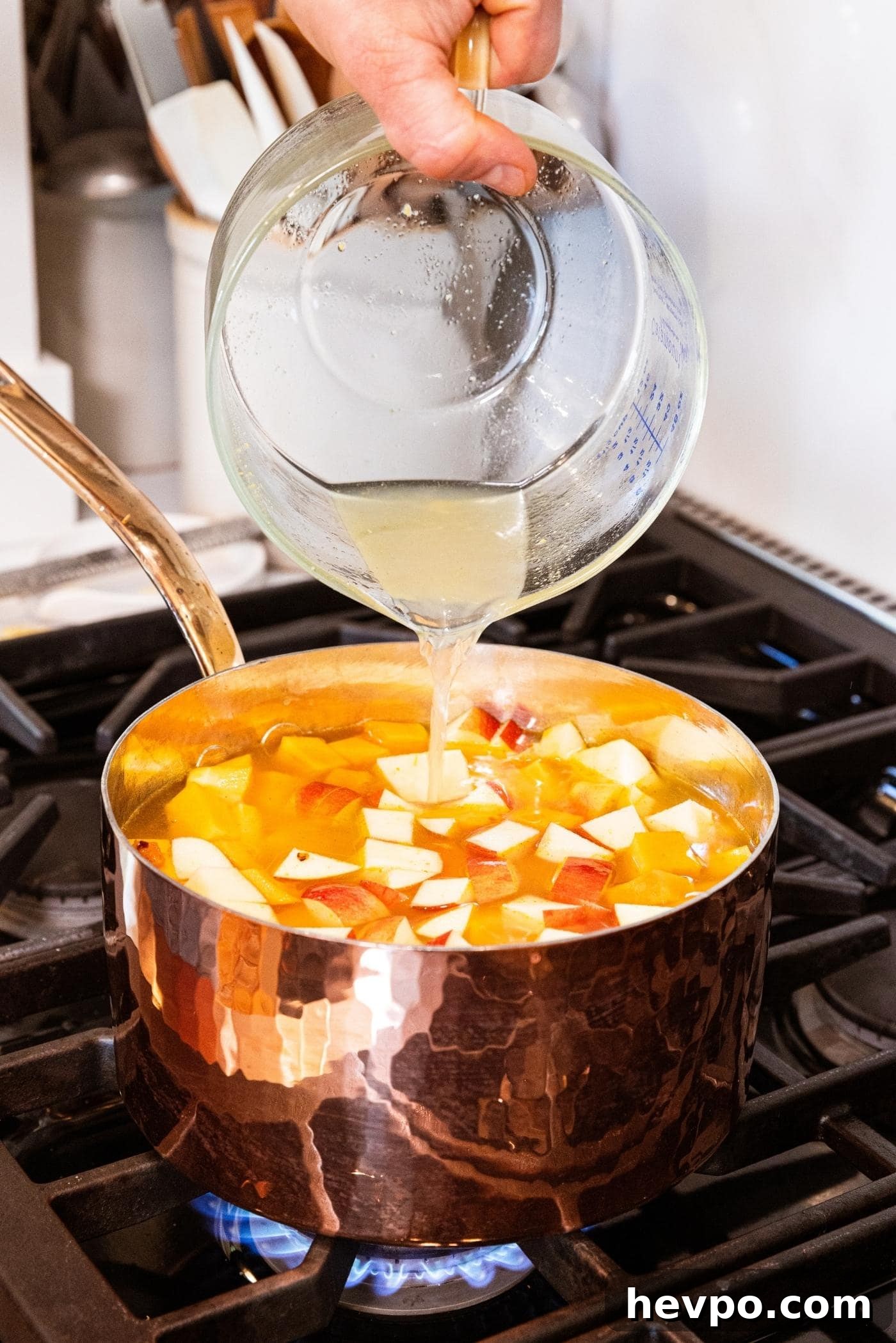 Hand pouring chicken stock from glass measuring cup into copper pot filled with diced butternut squash and apple.