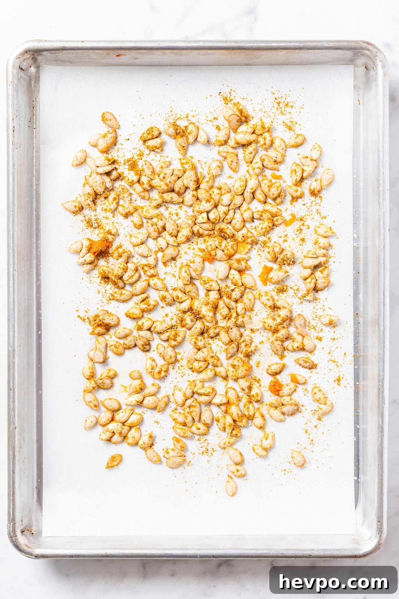 Metal baking pan with piece of white parchment paper with squash seeds spread out in even layer sprinkled with curry powder.