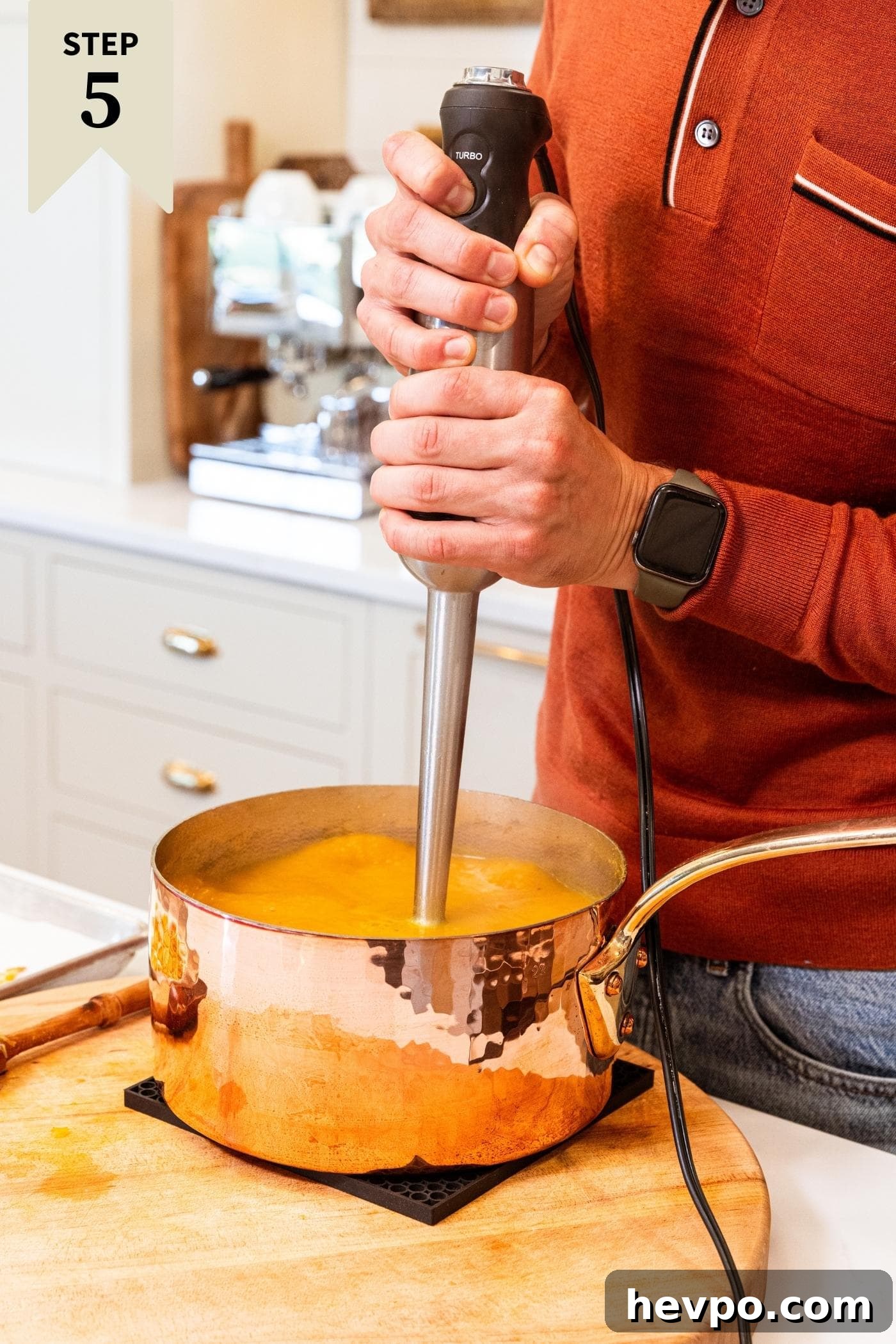 Two hands holding immersion blender blending butternut squash soup sitting in copper pot on wooden cutting board on white countertop.