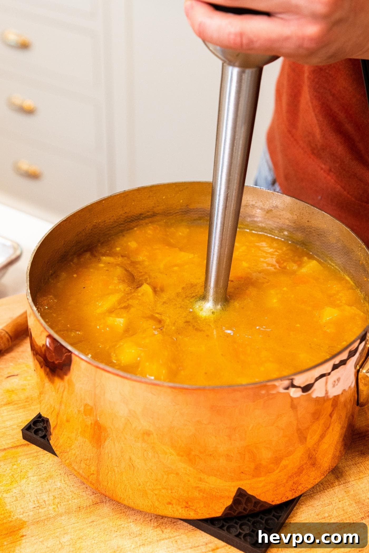 Copper pot sitting on black cooling hotpad on wooden cutting board with immersion blender in middle of pot blending the soup in pot together.