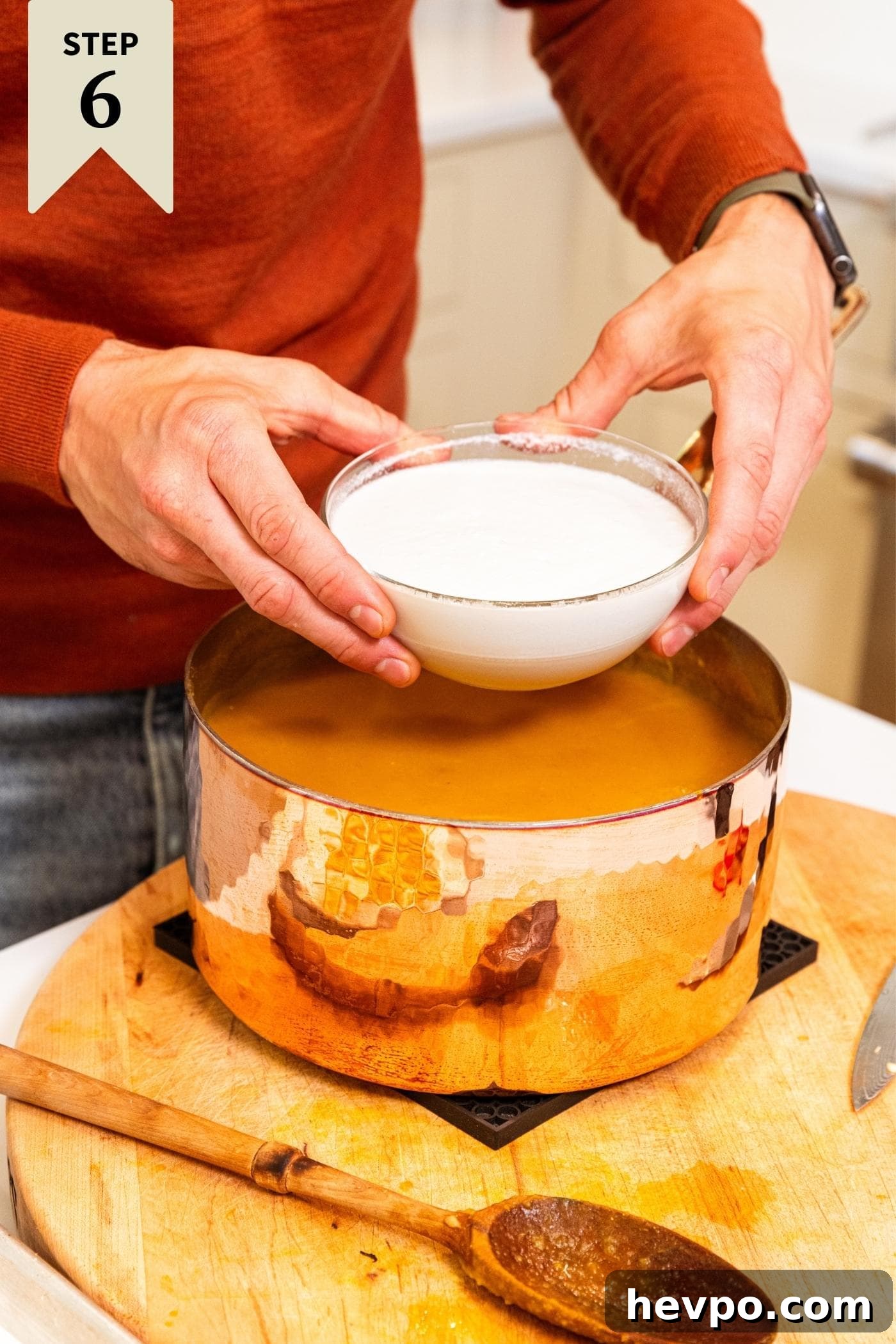 Two hands holding glass bowl filled with coconut milk pouring into a copper pot filled with butternut squash soup on wood cutting board.