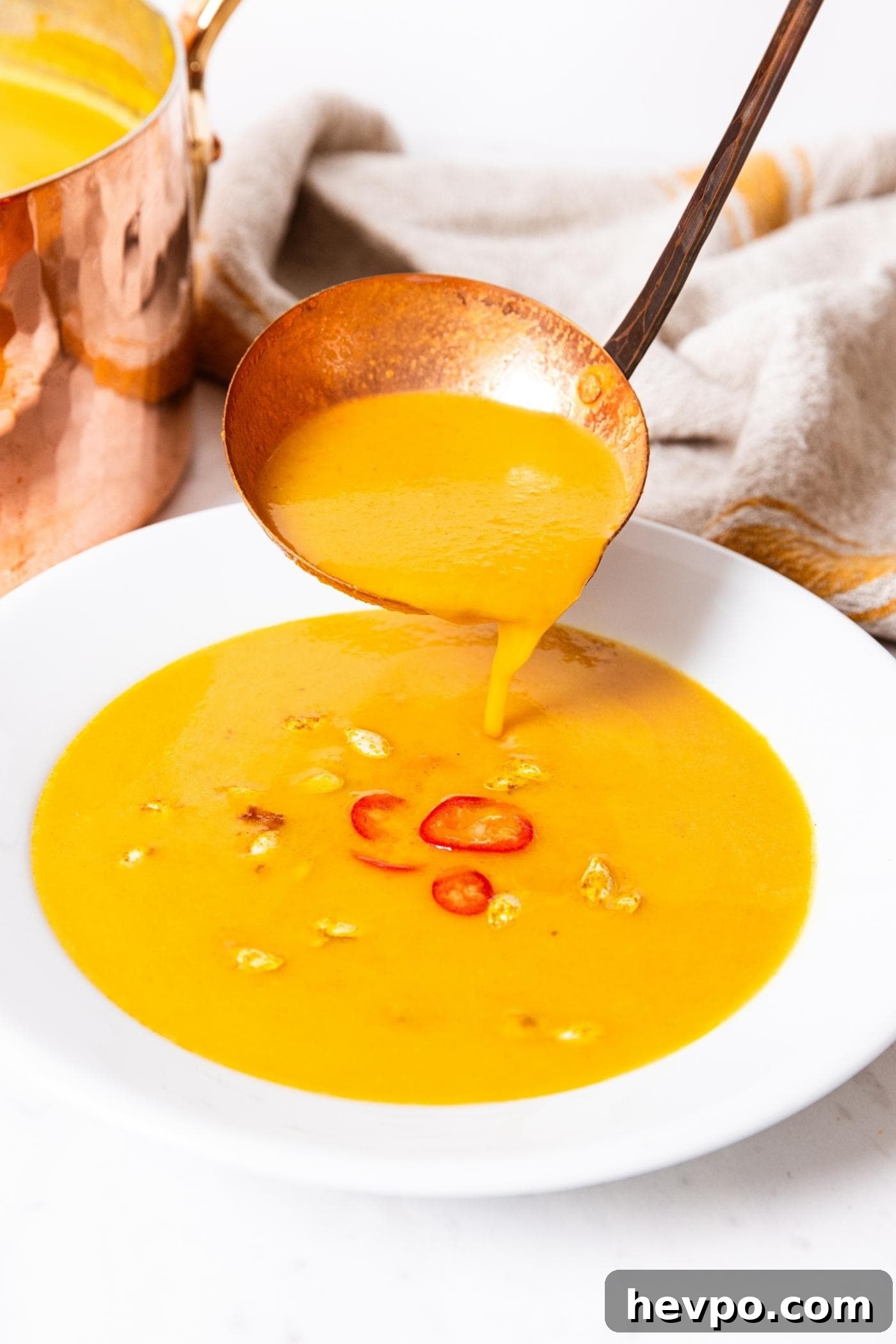 Copper ladle pouring serving of soup into a white bowl on marble surface with a few pieces of red pepper and squash seeds floating on top.