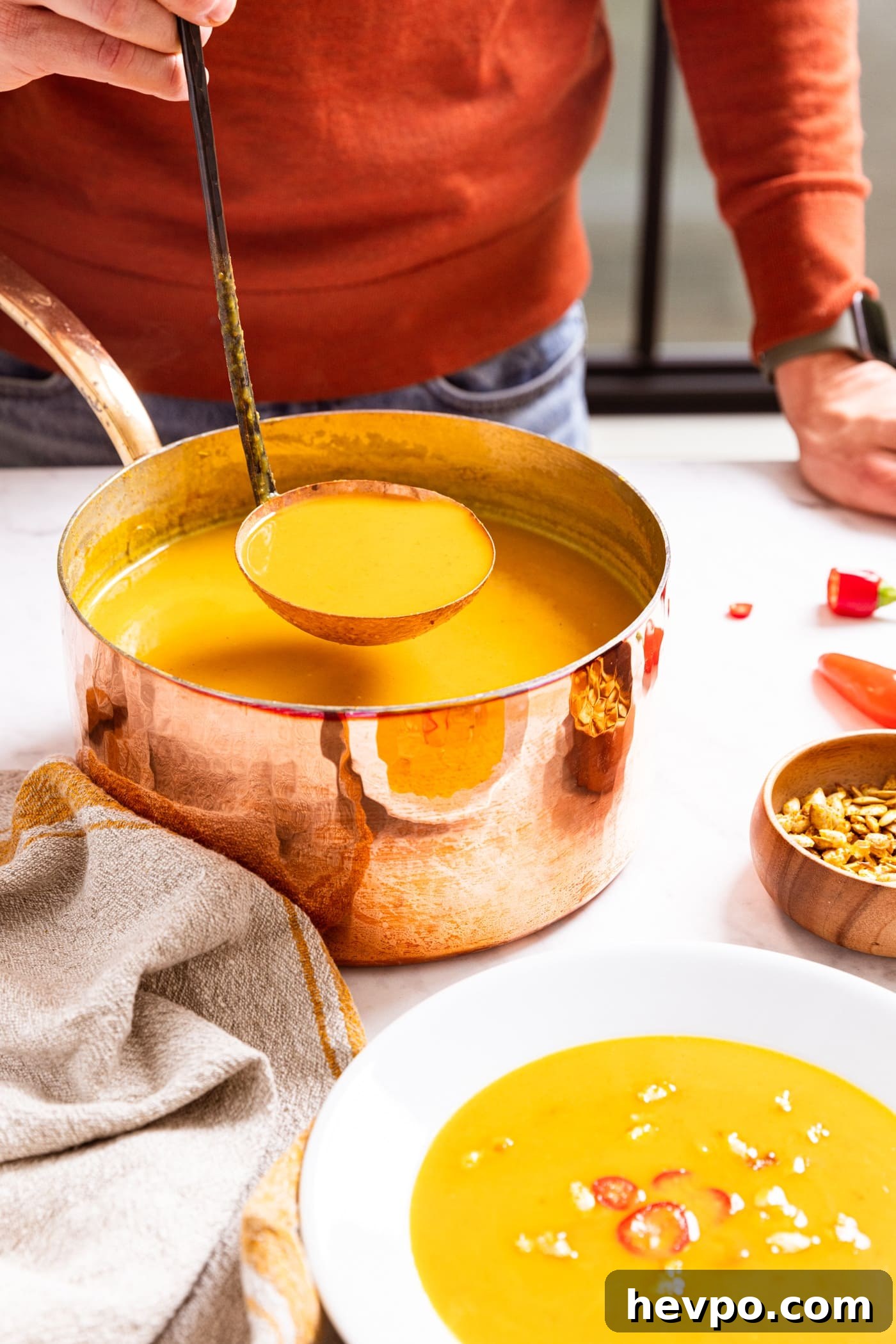 Hand holding copper colored ladle scooping out serving from copper stockpot sitting on marble surface with serving of soup already in white bowl.