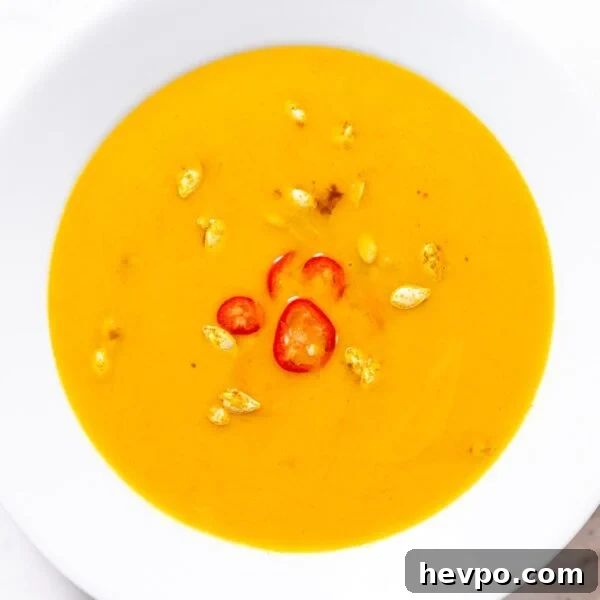 White bowl filled with curried butternut squash soup with toasted seeds and sliced red chiles on top all on white marble surface.