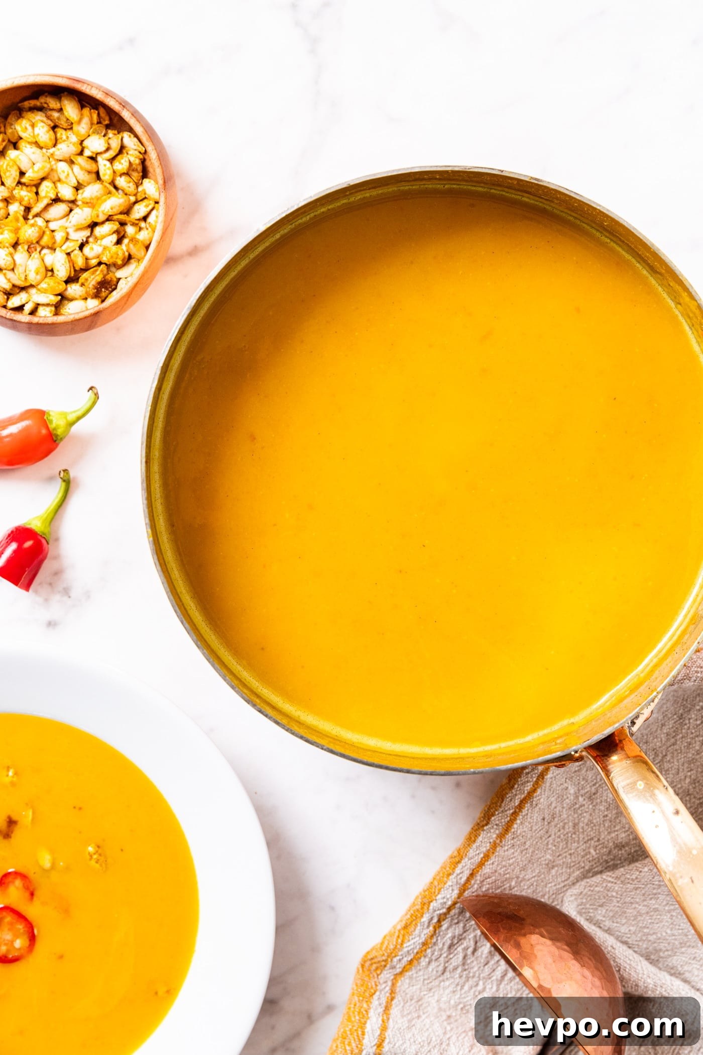 Copper pot filled with yellow colored squash soup sitting on marble surface with white bowl filled with soup and squash seeds in wooden bowl.