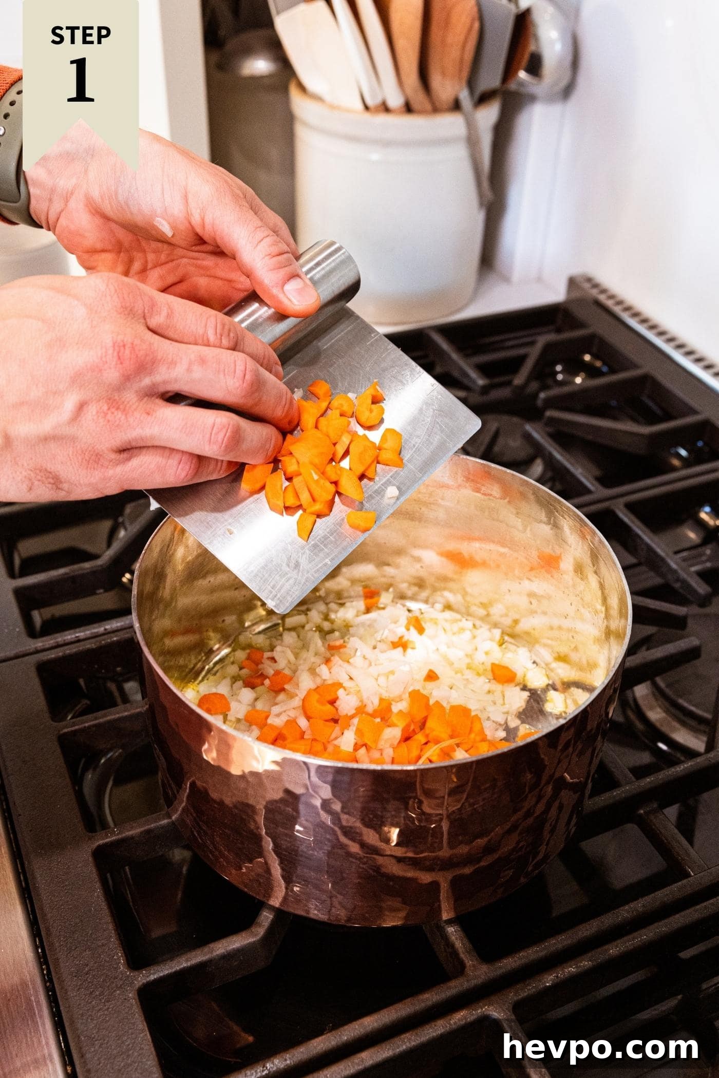 Hand holding metal bench scraper putting diced carrot pieces into copper pot on stove with more carrots and onion already heating.
