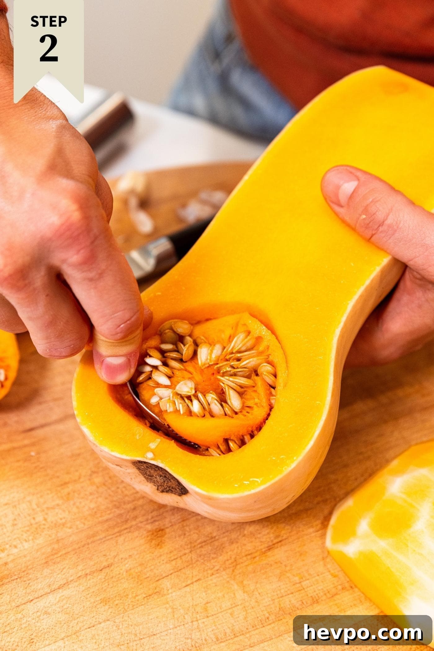 Hand using spoon to scoop out seeds from a butternut squash on a wood cutting board.