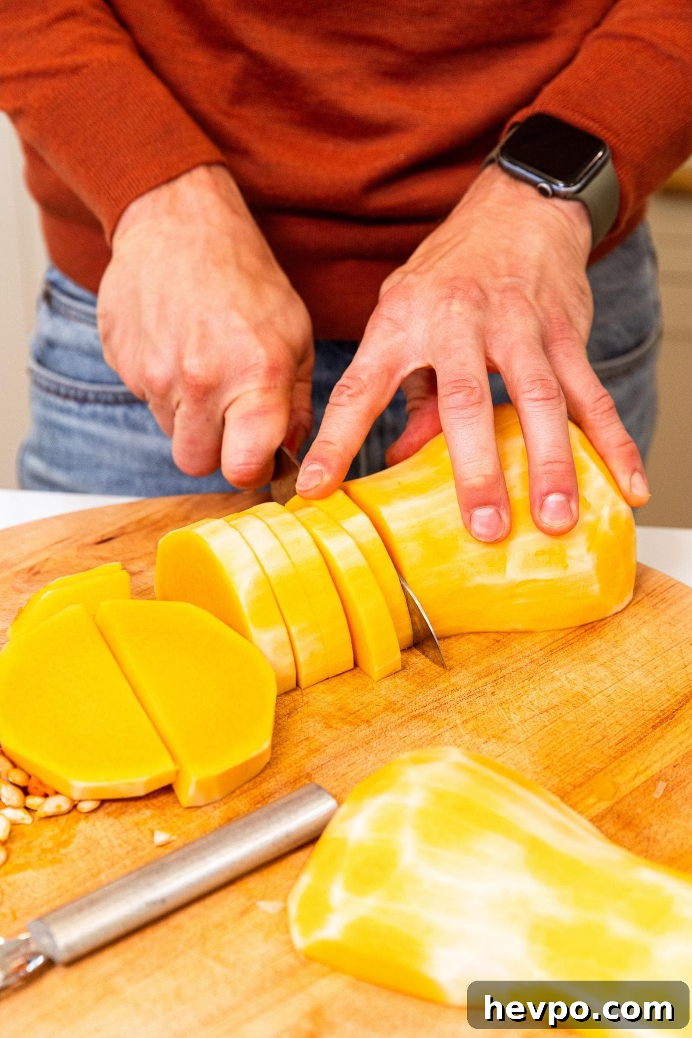 Hand slicing butternut squash into slices before dicing on wood cutting board.