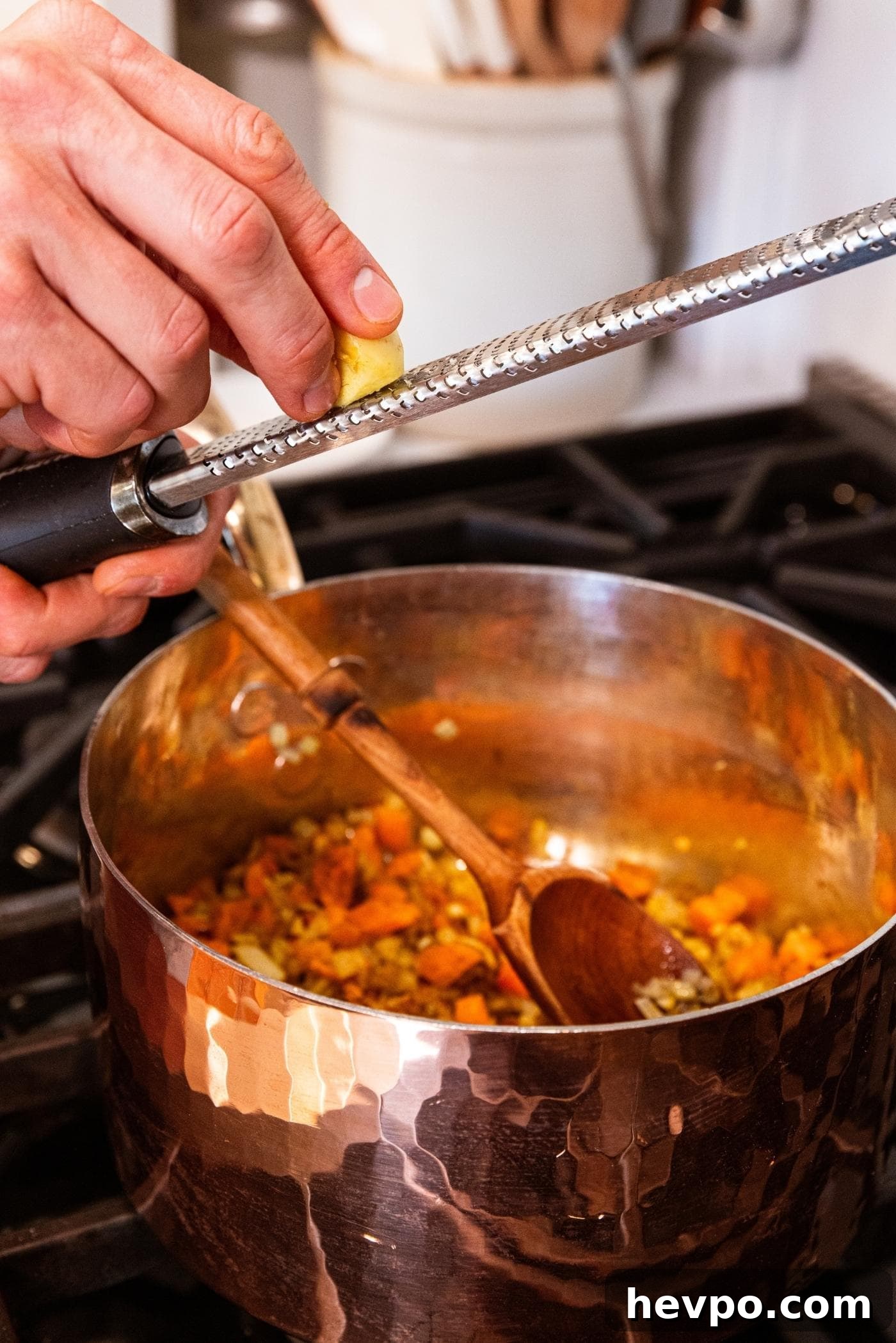 Hand using grater to grate fresh ginger into copper pot sitting on stove with spices and carrots and onion inside.