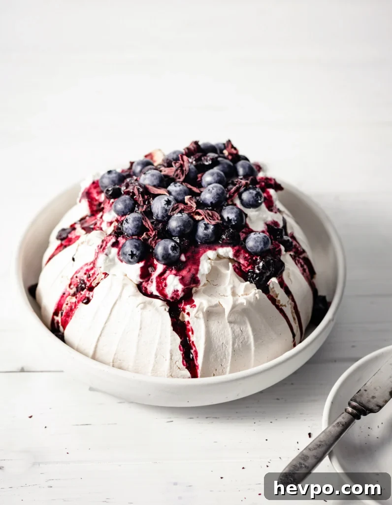 Baked meringue pavlova inside a large serving bowl, topped with whipped cream, hibiscus compote and blueberries, on a wood table.