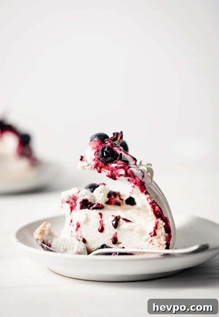 A slice of pavlova on a plate, topped with whipped cream, hibiscus compote and blueberries, on a wood table.