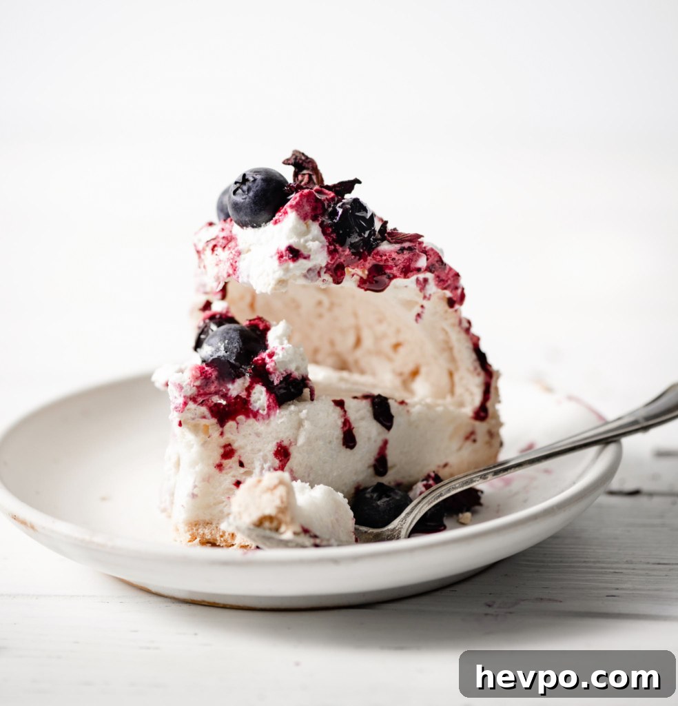 A slice of pavlova on a plate, topped with whipped cream, hibiscus compote and blueberries, on a wood table.