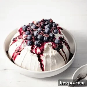 Baked meringue pavlova inside a large serving bowl, topped with whipped cream, hibiscus compote and blueberries, on a wood table.