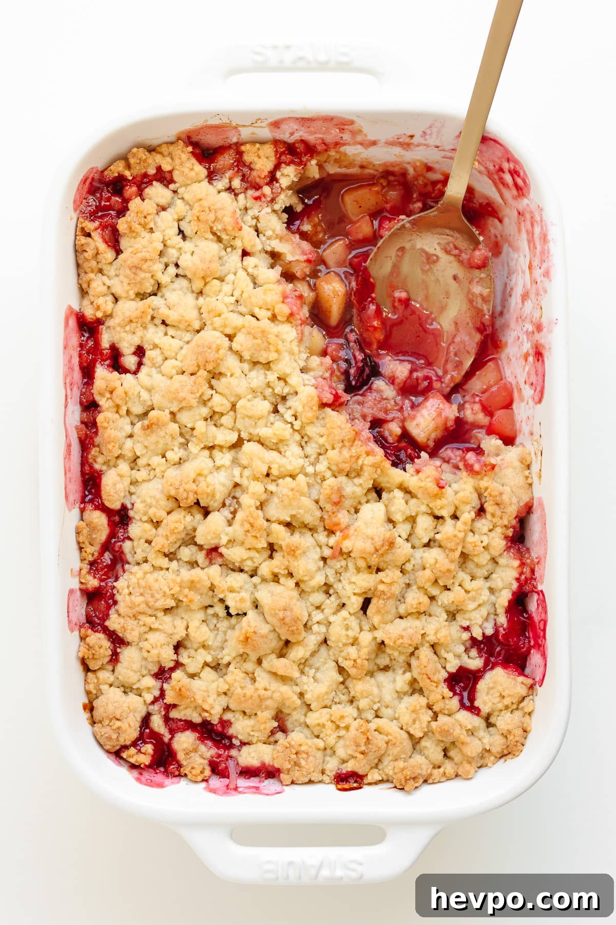 Plum apple crumble in a white baking dish with a serving spoon sitting in a corner where a portion has been scooped out.