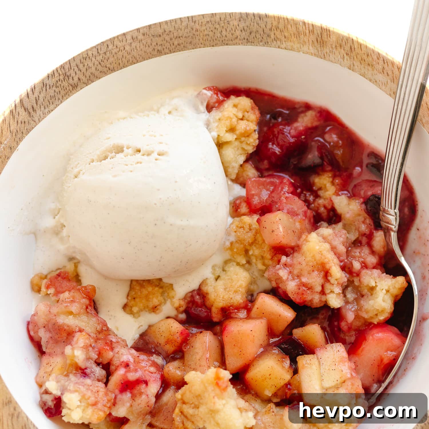 Bowl of plum and apple crumble with a spoon and scoop of vanilla ice cream melting on top.