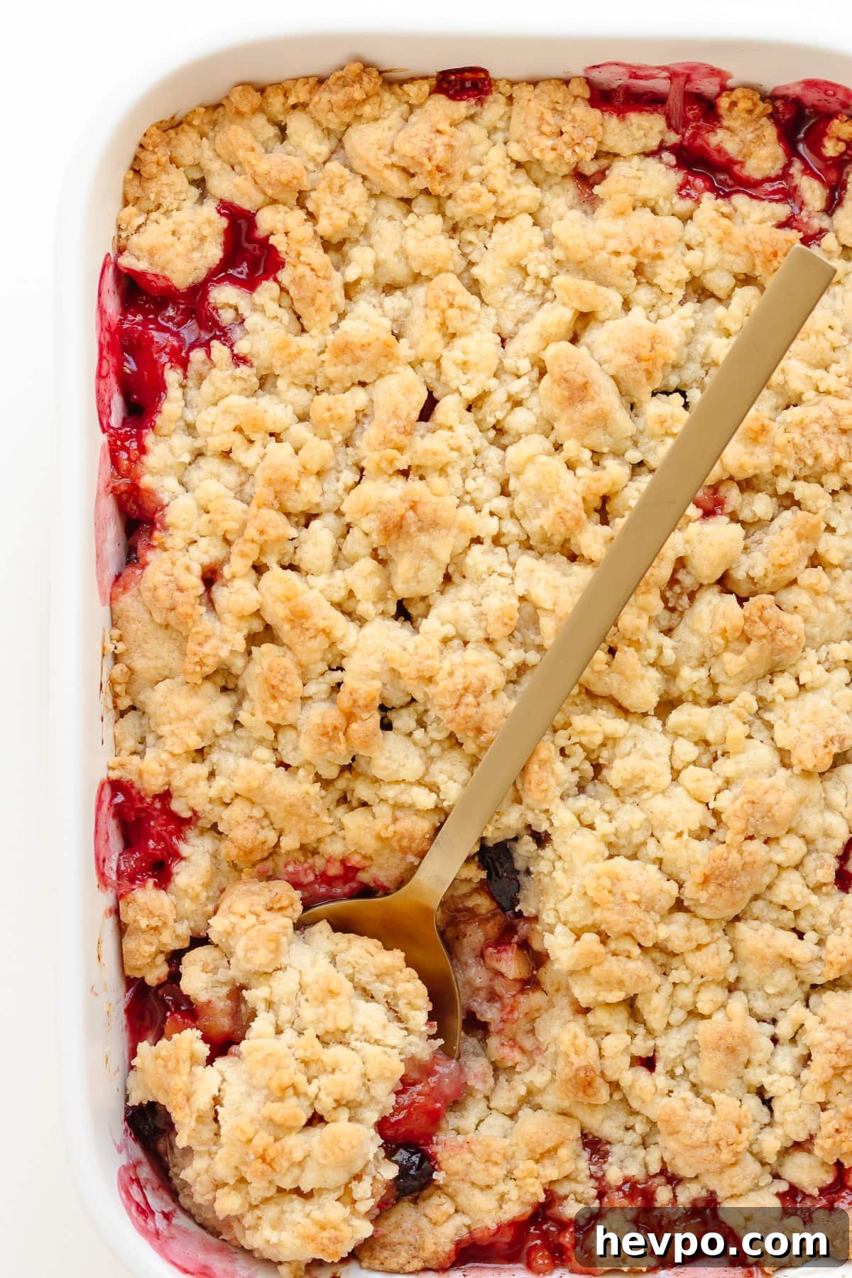 Apple and plum crumble in a white baking dish with gold colored serving spoon.