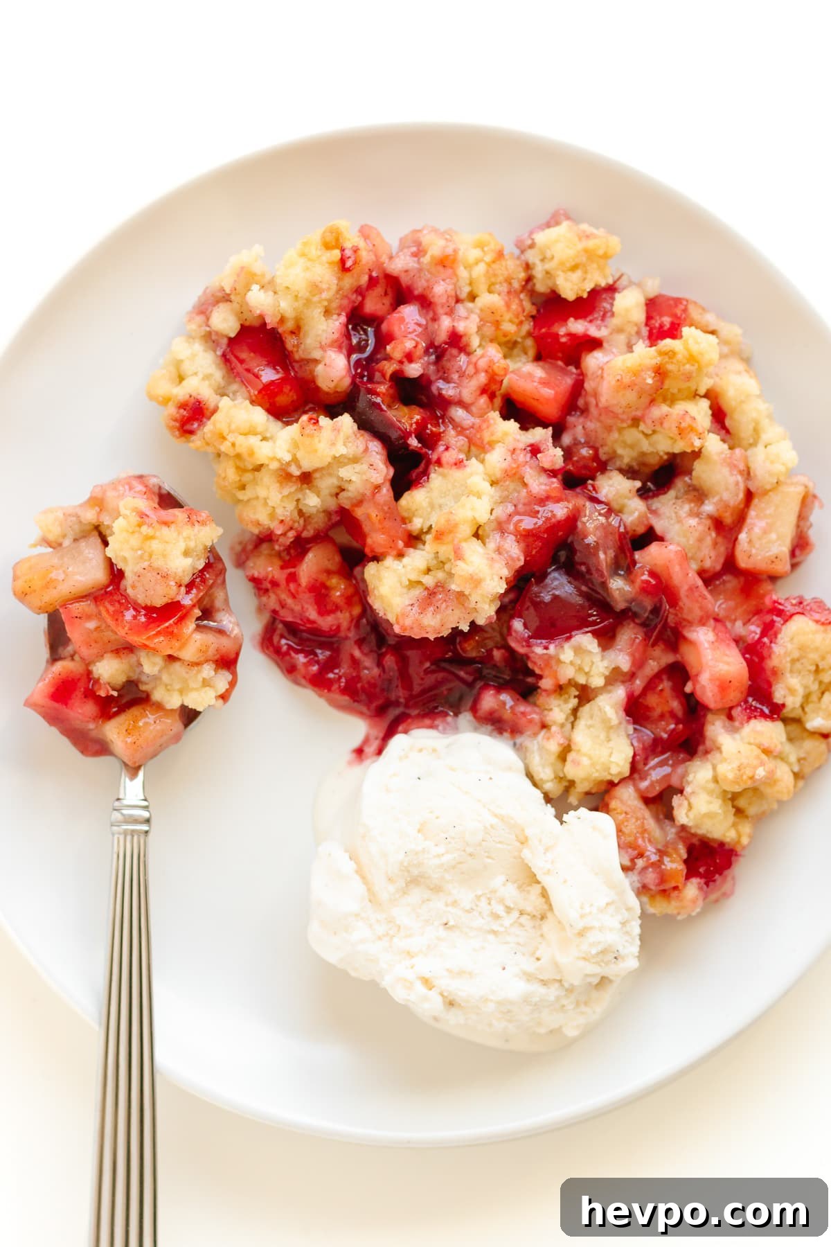 Plum and apple crumble on a white plate with a scoop of vanilla ice cream and spoon.