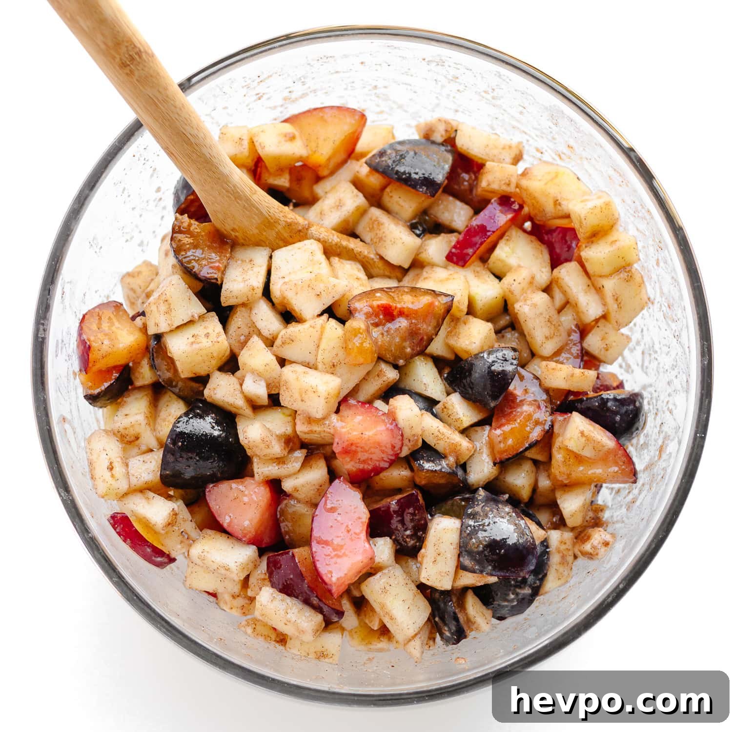 Plum and apple fruit mixture for crumble being mixed in a glass bowl with a wooden spoon.