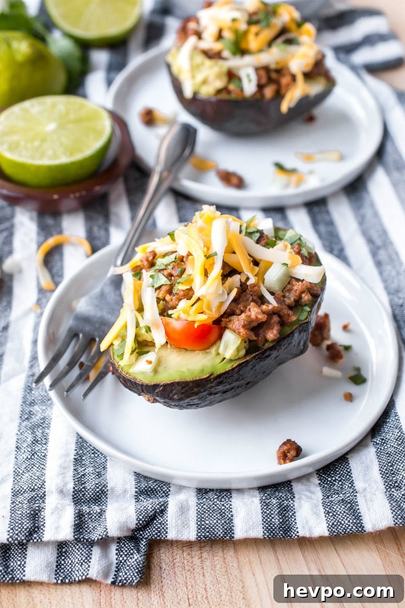 half an avocado stuffed with taco filling, sitting on a white plate with a fork. Lime in the background. Sitting on a blue and white striped napkin.