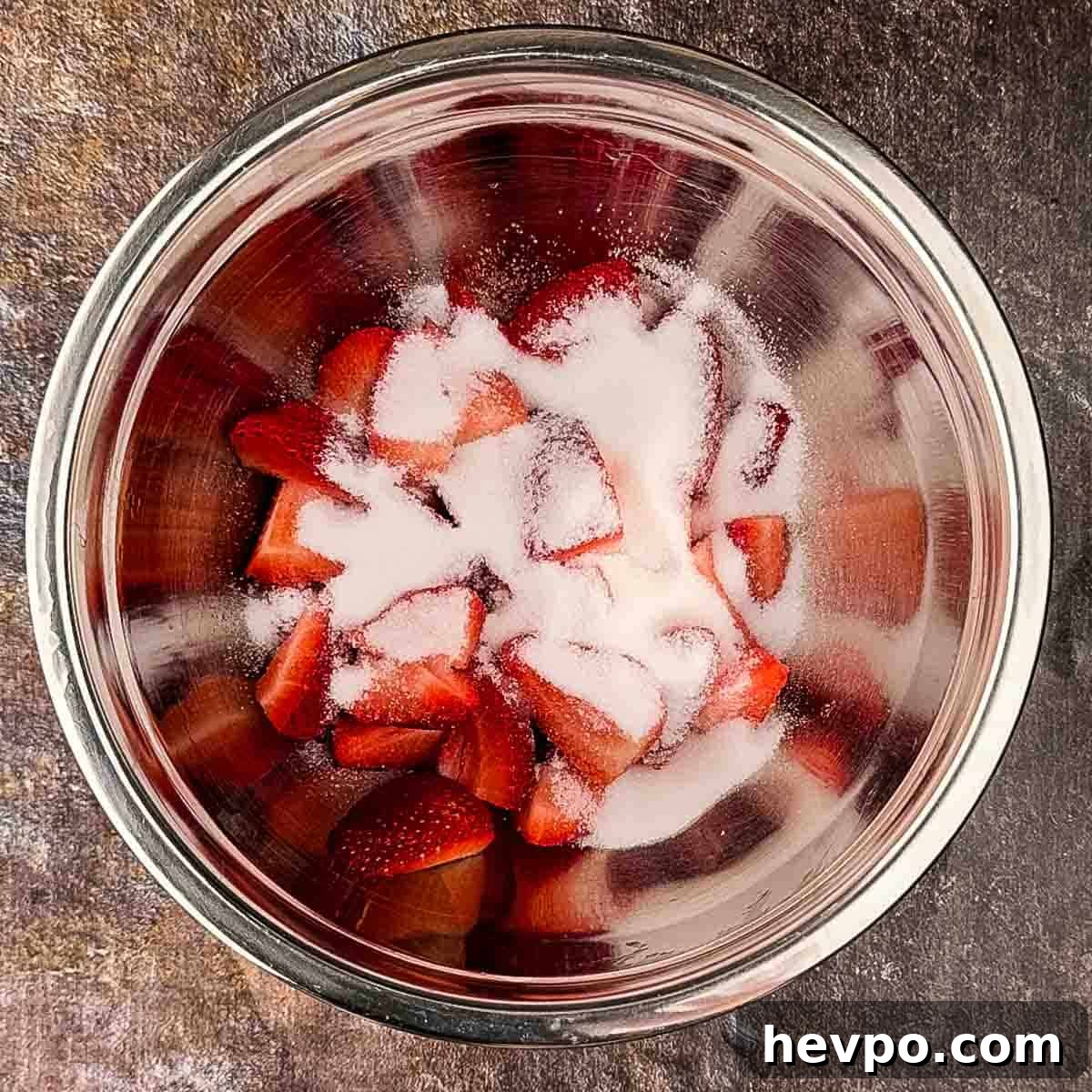 Sliced strawberries and sugar in a steel mixing bowl.