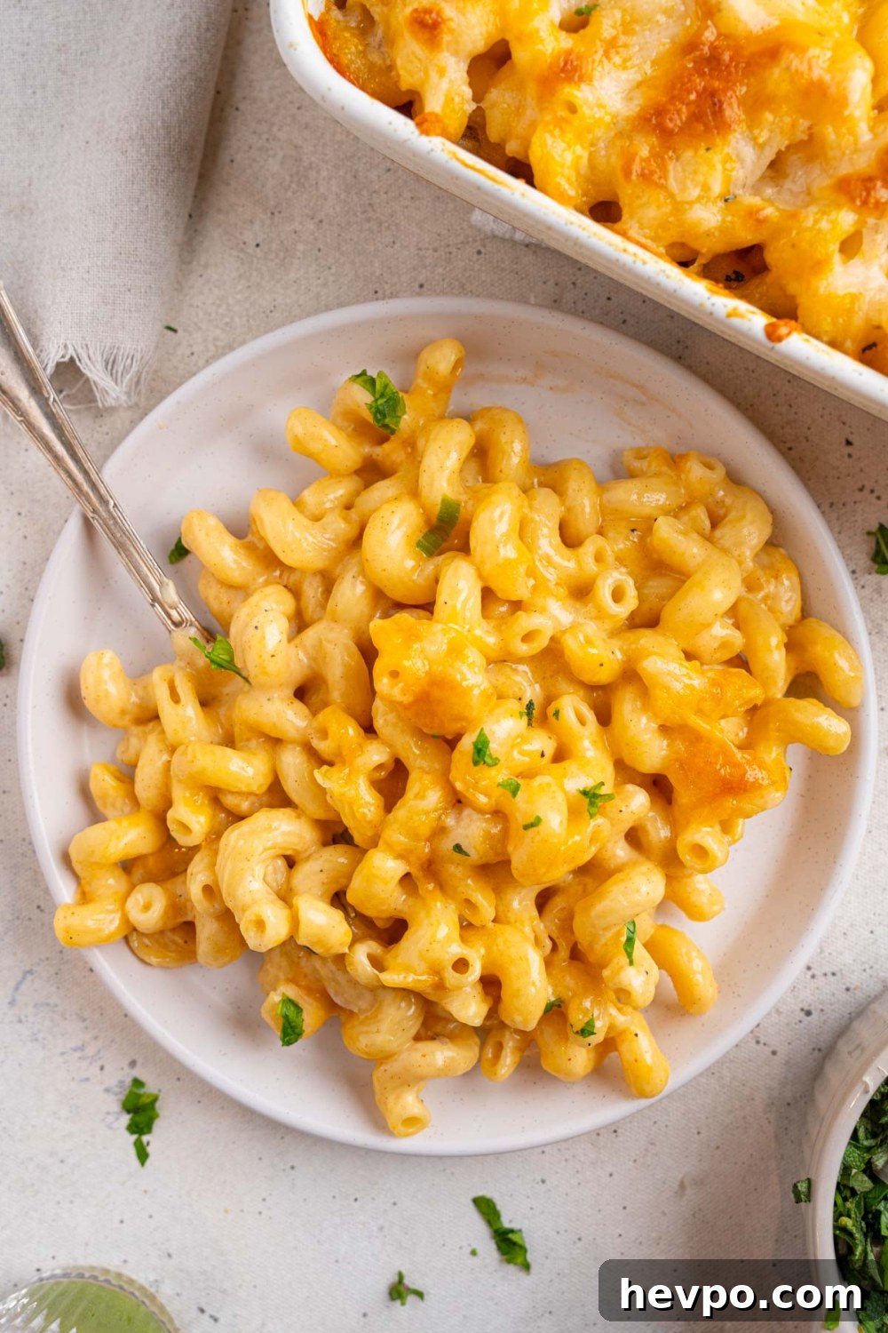 Tini’s Viral TikTok Mac and Cheese Recipe 3 A white ceramic plate with Tini's mac and cheese garnished with fresh parsley. There is a fork inserted in the plate and the plate is on a white counter with a baking dish of mac and cheese.