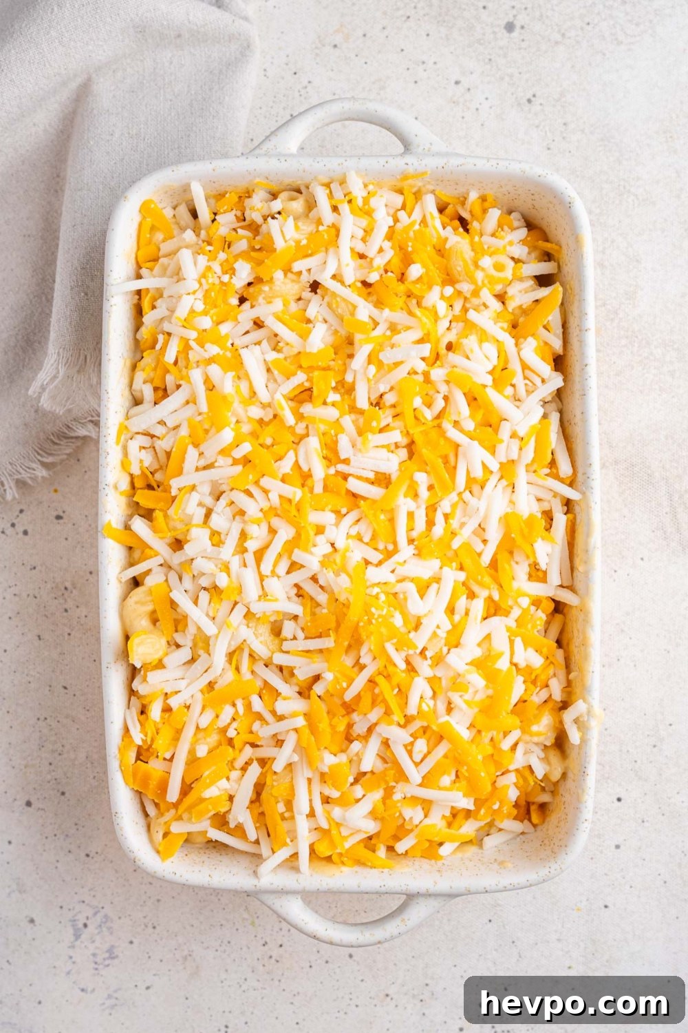 Tini’s Viral TikTok Mac and Cheese Recipe 9 A white ceramic baking dish with uncooked mac and cheese topped with a shredded cheese blend. The dish is on a white counter with a white cloth napkin.