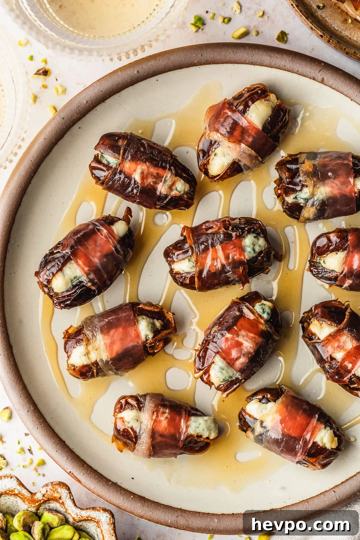 Prosciutto-Wrapped Dates with 3 Simple Ingredients 12 A stoneware plate of blue cheese stuffed prosciutto wrapped dates with honey on a tan counter next to a beige linen, brown bowl of pistachios, and glasses of white wine.