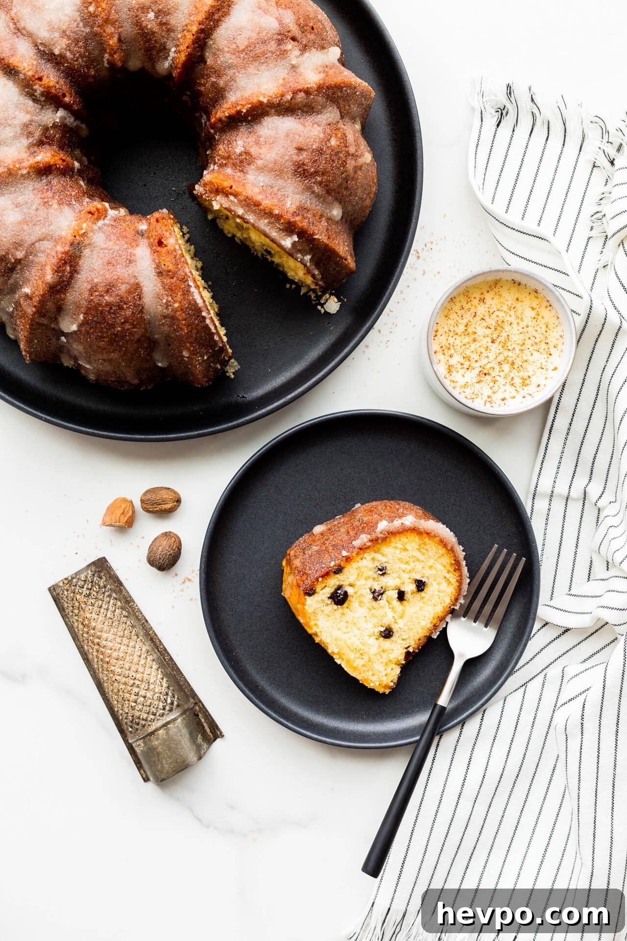 A slice of eggnog bundt cake with dried currants, served on black plate with a cup of eggnog and freshly grated nutmeg.