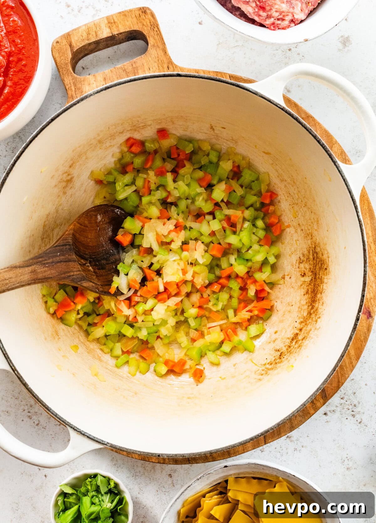 carrots, onion, and celery being stirred in a large pot with other lasagna soup ingredients in bowls surrounding the pot.