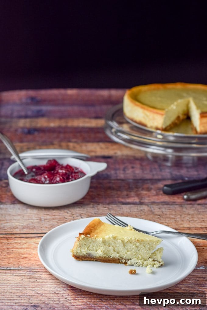A piece of cheesecake on a plate with cherry sauce in a bowl and the cake on the pedestal