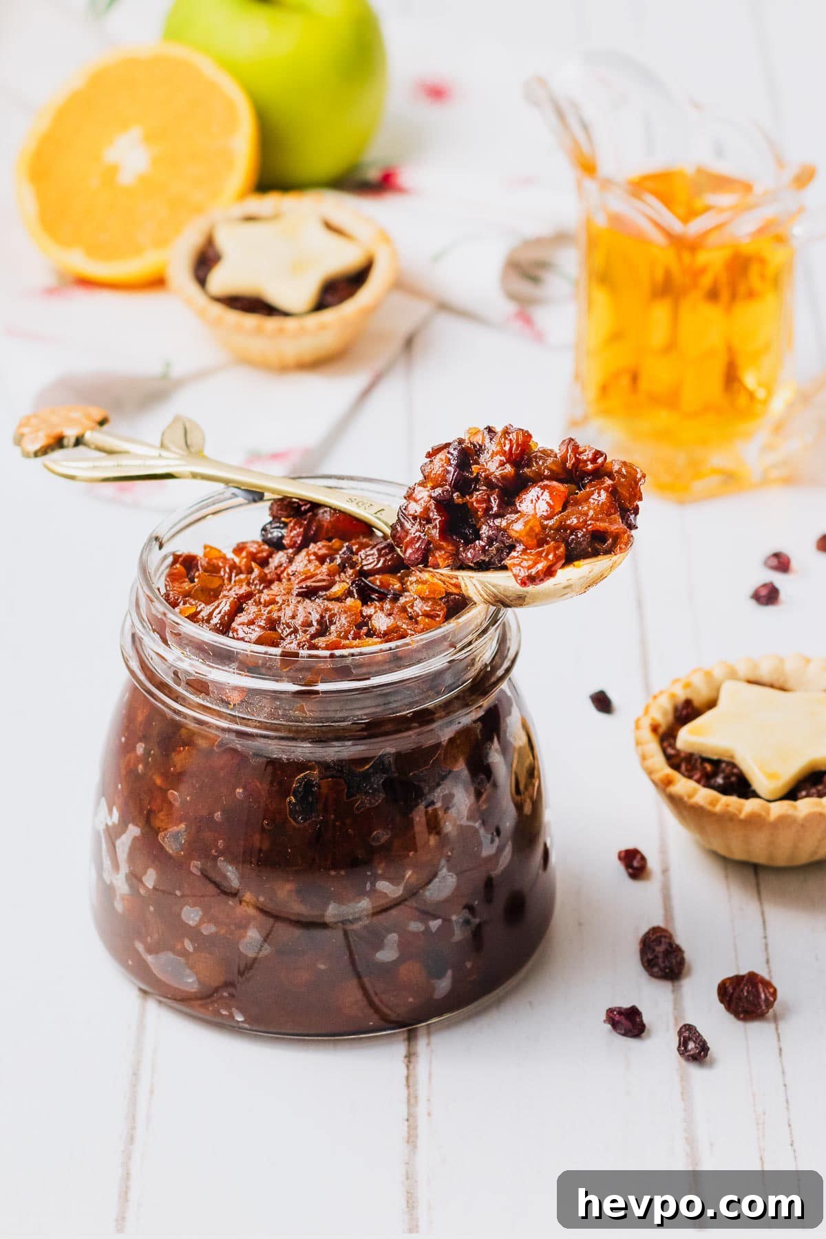 A large glass jar of fruit mince, on a white wooden background, with fruit mince pies, an apple, an orange and a glass jug of brandy, text overlay reads "Christmas fruit mince recipe".
