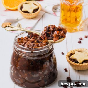 A large glass jar of fruit mince, on a white wooden background, with fruit mince pies and a glass jug of brandy in the background.