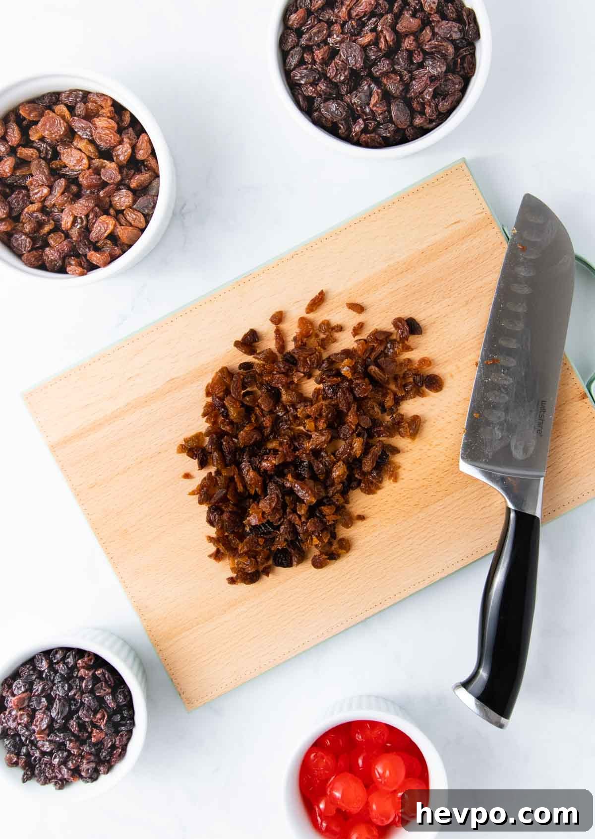 Chopped raisins on a wooden chopping board with a large kitchen knife and other dried fruit in small white bowls.