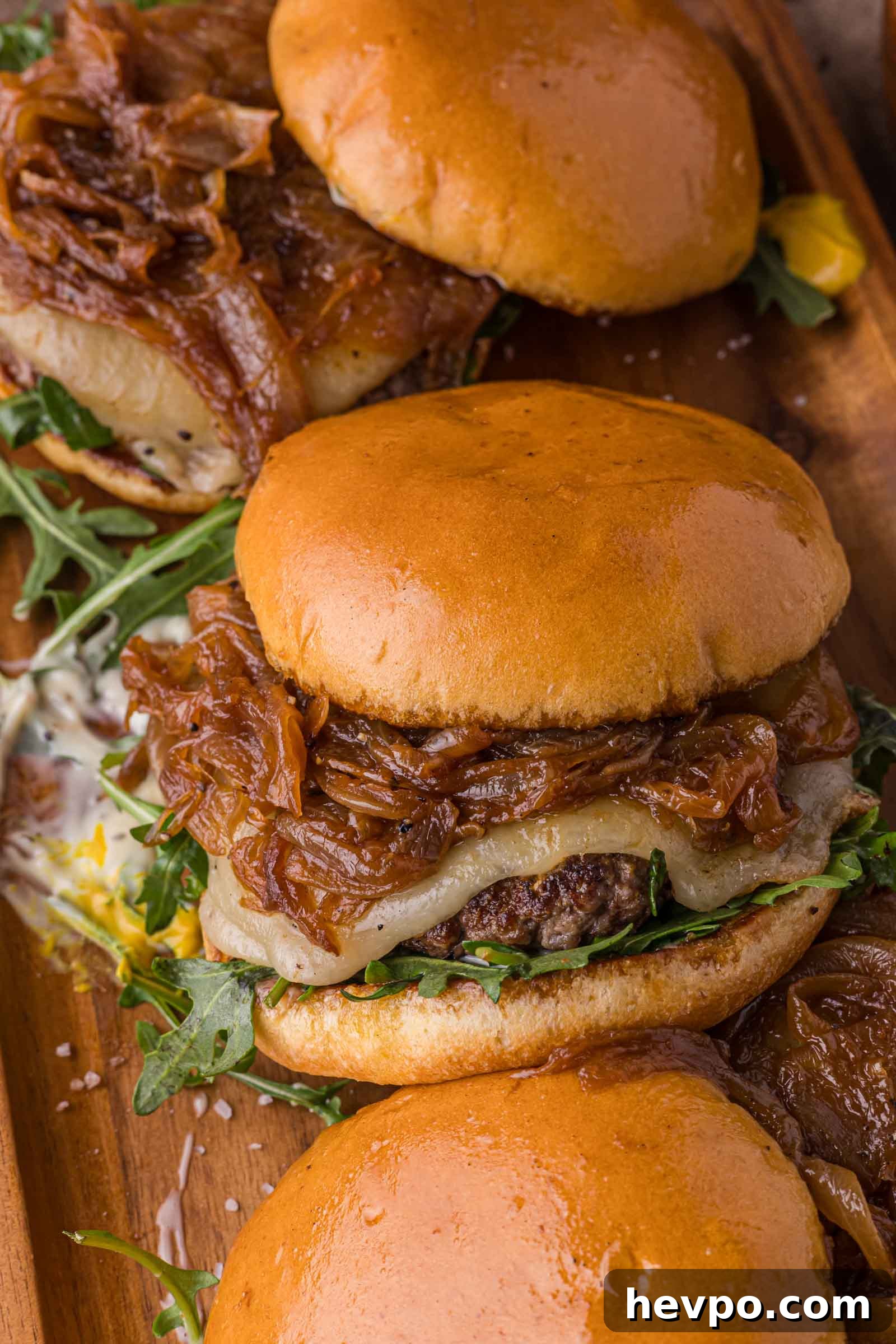 Three cheeseburgers on a wooden board, topped with caramelized onions, arugula, and melted cheese on toasted buns.