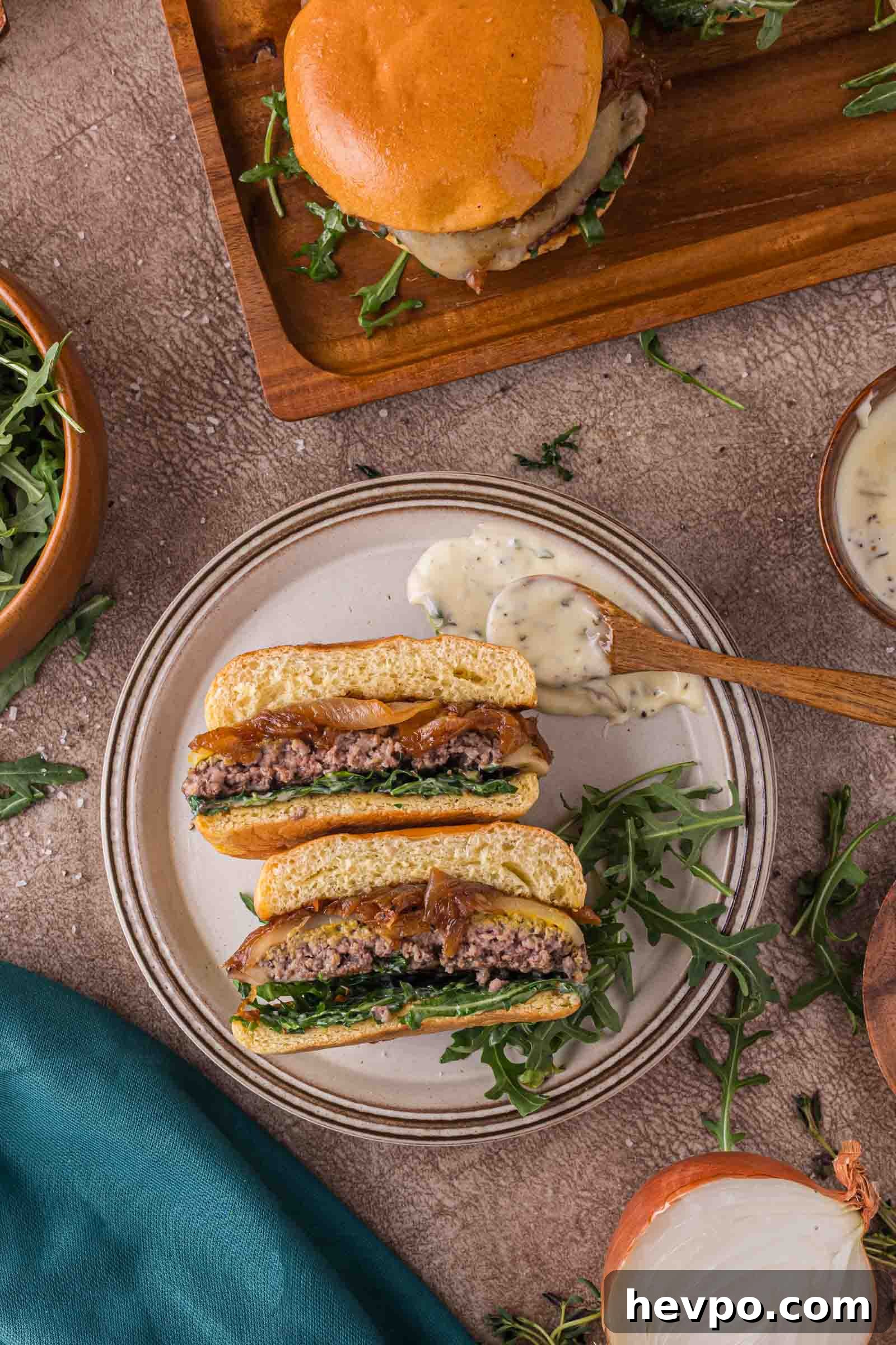 A sliced burger with arugula and onions on a plate, served with creamy aioli; another whole burger and bowls of arugula and sauce are nearby.