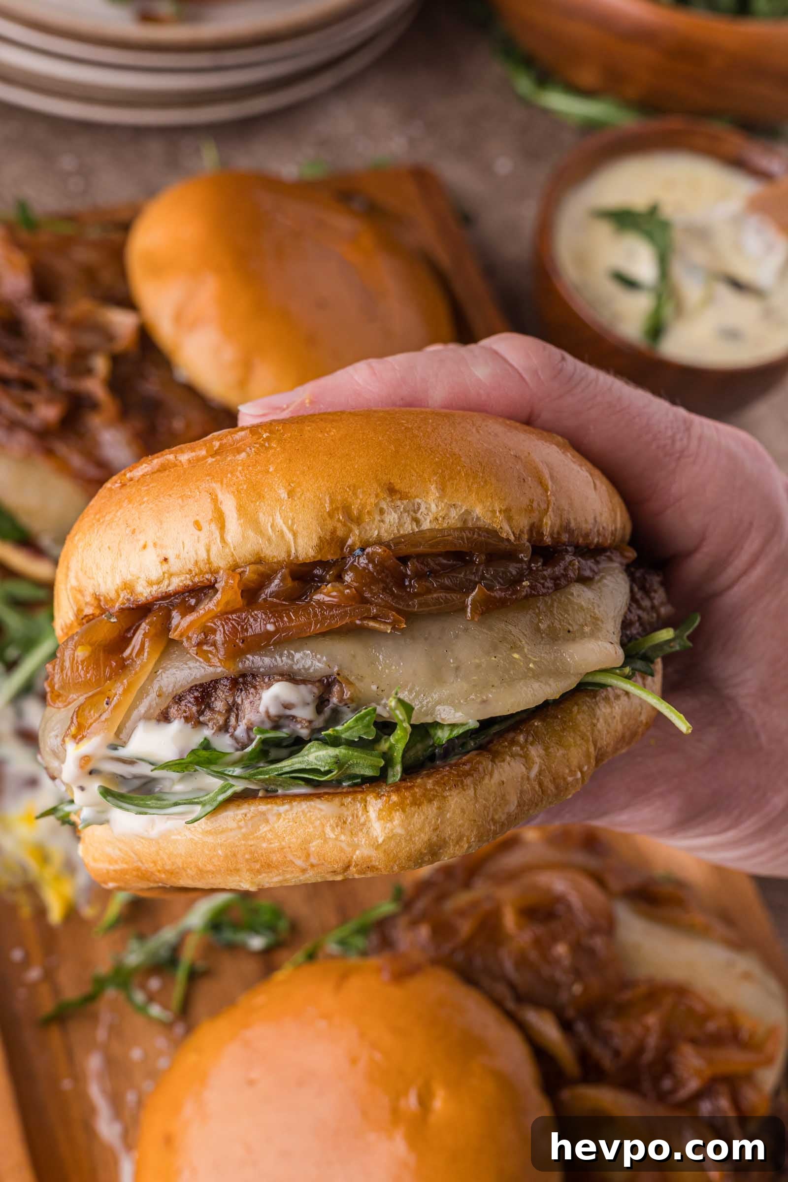 A hand holds a burger with caramelized onions, melted cheese, arugula, and sauce in a shiny bun. Other burgers and a bowl of sauce are visible in the background.