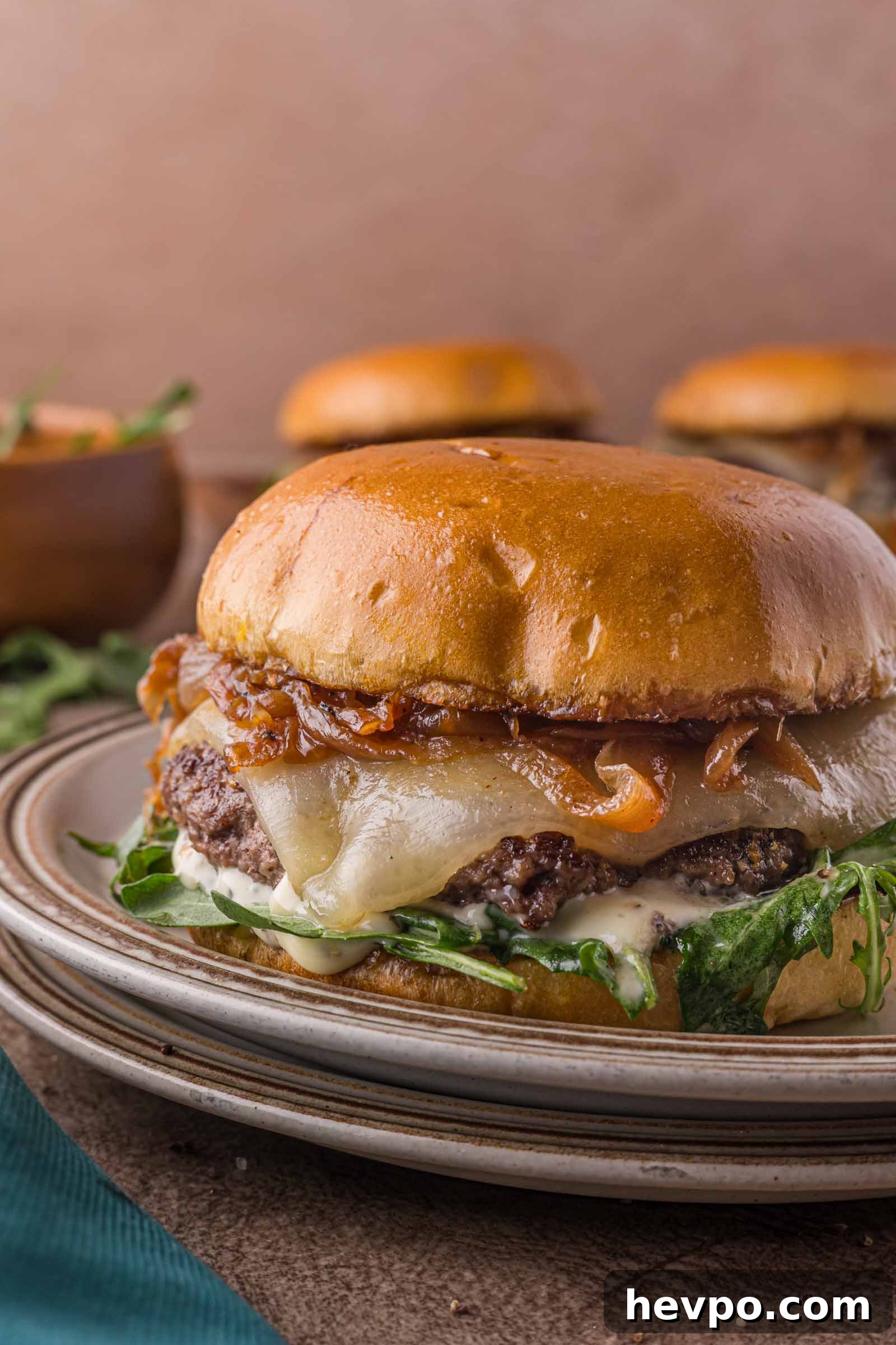 A cheeseburger with melted cheese, caramelized onions, greens, and sauce on a toasted bun sits on a plate.