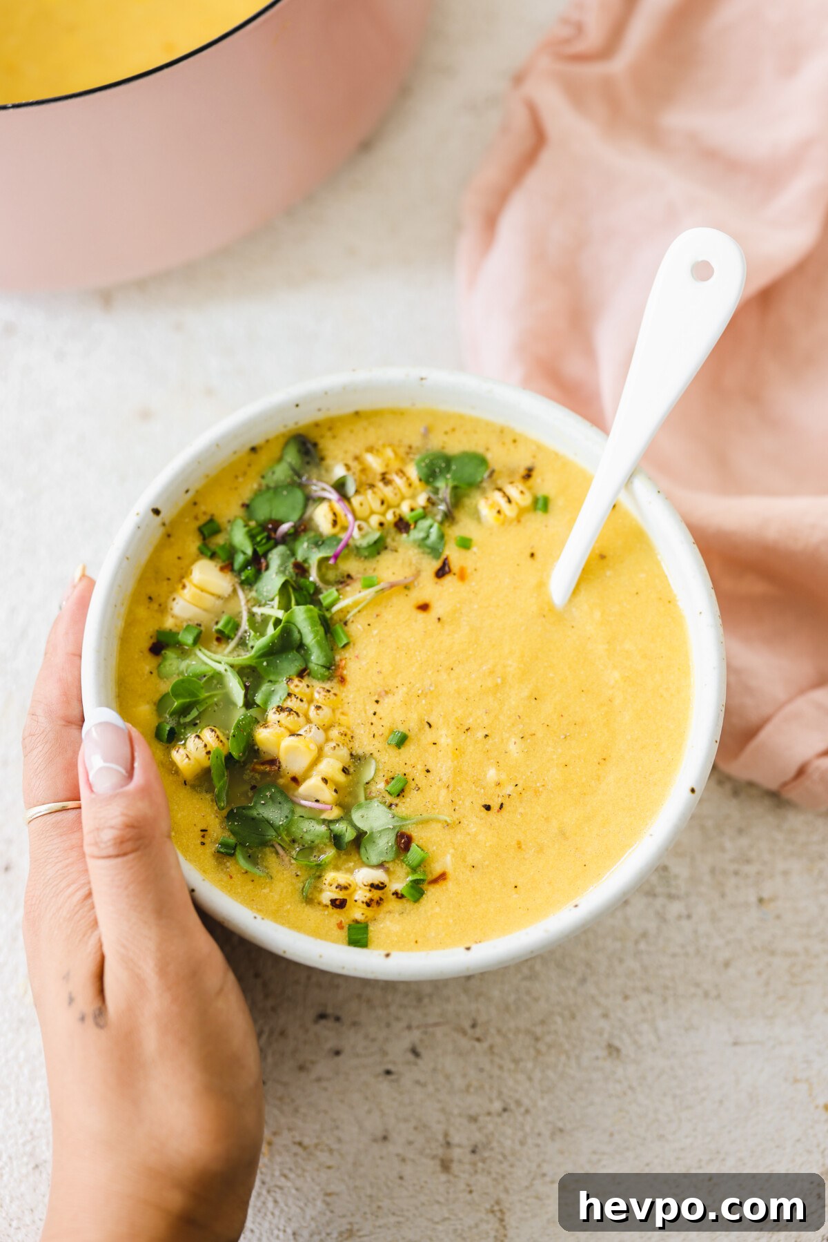 a hand holding a bowl of creamy vegan corn chowder topped with charred corn and microgreens