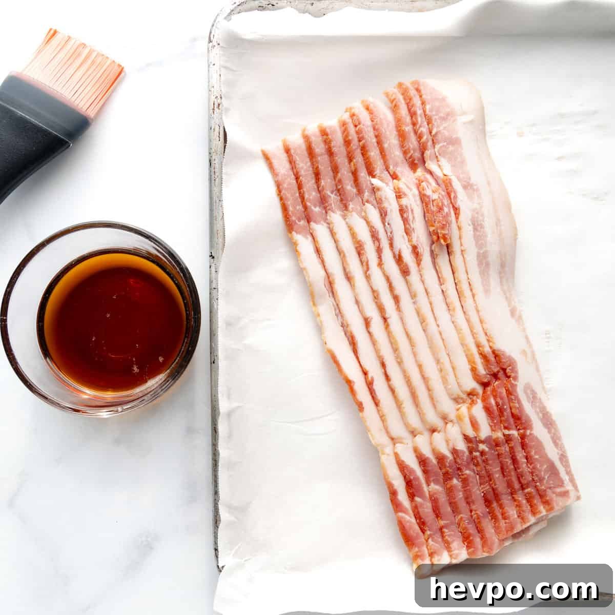Maple Bacon Recipe with Just 2 Ingredients 3 Slices of raw bacon on a lined baking sheet next to a glass cup filled with a dark liquid and a basting brush.